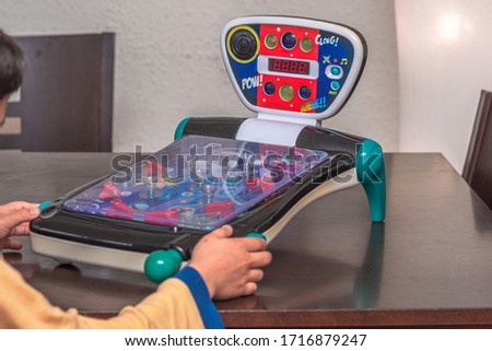 Mini pinball toy machine on a brown table. In photo one can see a couple of child´s hands on the pinball machine ready to play. Indoor photo. Artificial lighting Royalty-Free Stock Photo #1716879247