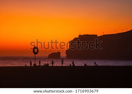 Nazare, Portugal: Amazing orange sunset over Atlantic Ocean and people relaxing on the beach. Lighthouse and Fort of Sao Miguel visible on the high cliff.