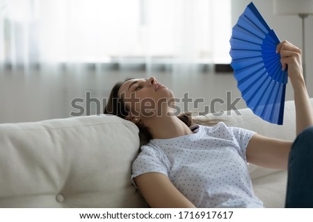Exhausted young woman sit rest on sofa at home use waver suffering from hot weather indoors, overheated unwell millennial female breathe fresh air, wave with hand fan, having heatstroke Royalty-Free Stock Photo #1716917617