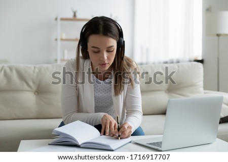 Concentrated millennial girl in headphones sit on couch at home make notes studying online on laptop, focused young woman watch webinar or Internet course on computer, distant education concept Royalty-Free Stock Photo #1716917707