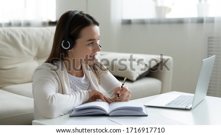 Happy millennial girl in headphones make notes watch webinar on laptop, smiling young woman in headset taking online course or web training, study on computer at home, distant education concept Royalty-Free Stock Photo #1716917710