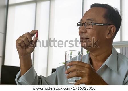 Senior asian man taking red pills with water glass at home.