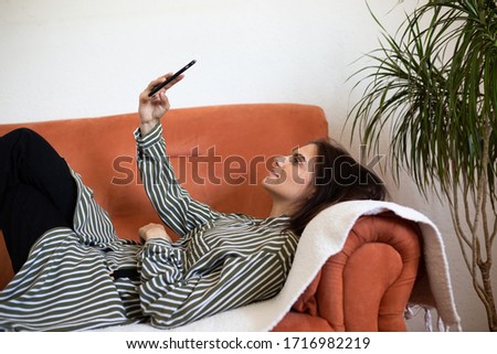 Fashionable young Caucasian woman laying on sofa at home holding mobile phone. She is texting on smartphone and taking selfie. 
