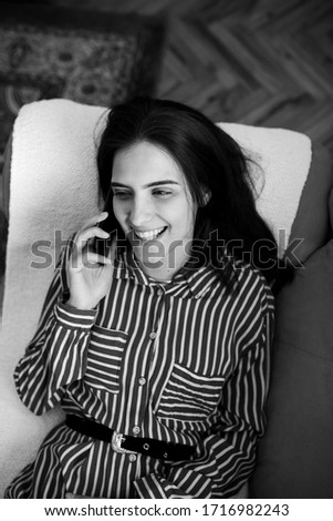Fashionable young Caucasian woman laying on sofa at home holding mobile phone and talking with friend. She is smiling and enjoying conversation. Black and white photo.