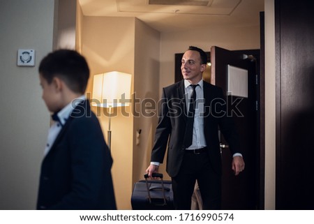 Father and son in formal wear entering a hotel room, the father is carrying a large suitcase