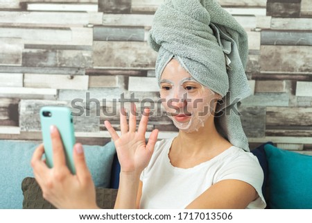 A girl in a cosmetic face mask and a towel on her head, with a phone in her hand, communicates via video communication. Makes homemade beauty treatments. Close up photo