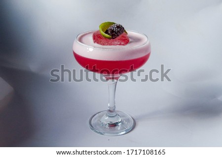 A clover club drink. It is pink and has a white foam on top, served on a cocktail glass with a chunk of ice and a fresh blackberry on top.