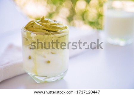 Home made Dalgona whipped macha in a cup on white table with a cup of milk and green nature blurred background.