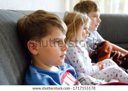 Cute little toddler girl and two school kid boys watching cartoons or movie on tv. Three happy healthy children, siblings during coronavirus quarantine staying at home. Brothers and sister together.