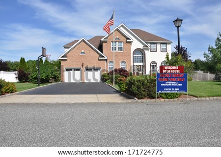 American Flag pole Welcome open house real estate sign curbside front yard Suburban McMansion style brick home Landscaped sunny residential neighborhood USA blue sky clouds