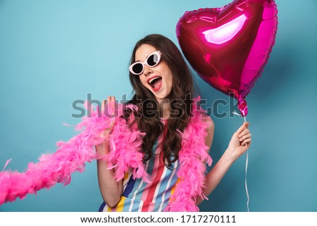 Photo of delighted young woman in sunglasses posing with balloon at camera isolated over blue wall