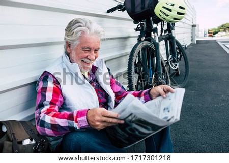 Happy senior man stops driving with electric bicycle and reads the newspaper sitting on the sidewalk. Active retiree and healthy lifestyle concept