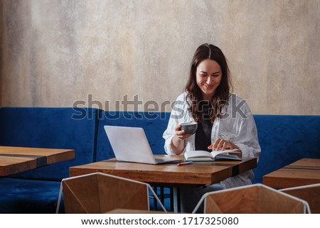 A portrait of a young girl sitting in a cafe and working on a laptop, drinking coffee, keeping notes in a notebook very fun and easy