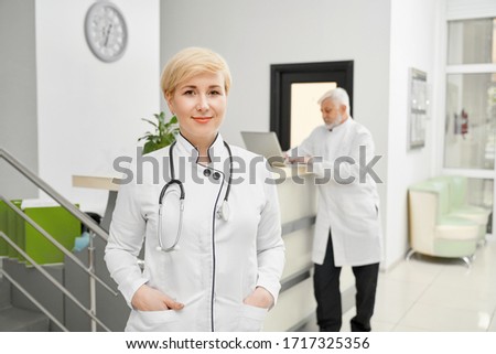 Professional therapist with stethoscope on neck posing looking at camera, eldery man using laptop on reception on background.