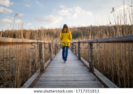 Behind a single Caucasian woman walking on a long wooden bridge near a beautiful reed-covered lake and deciduous forest during sunset. Young girl walking along the path alone towards unknown places. Royalty-Free Stock Photo #1717351813