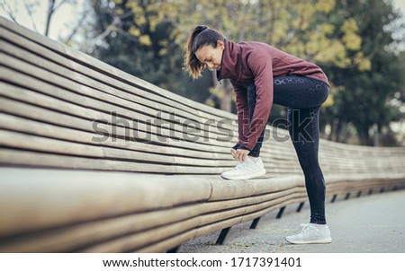 Sportswoman tying a shoe lace in a park. She is a runner. She is wearing garnet sweatsuit, black leggins and white trainers. She have the foot on a wood bank. Royalty-Free Stock Photo #1717391401