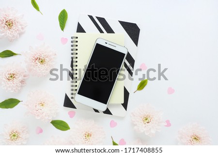 white smartphone with a black screen on yellow and black and white notepads and many pink flowers of chrysanthemums and green leaves on a white table. space for text.

