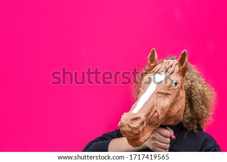 Portrait of blonde curly girl holding toy of horse's head on bright pink background. Trend photo without face in minimal style.