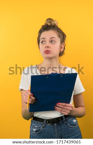 A young girl with her hair gathered in a bundle looks to the side, she ponders, holds a pencil and a notebook in her hand, in a white T-shirt on a yellow background, vertical.
