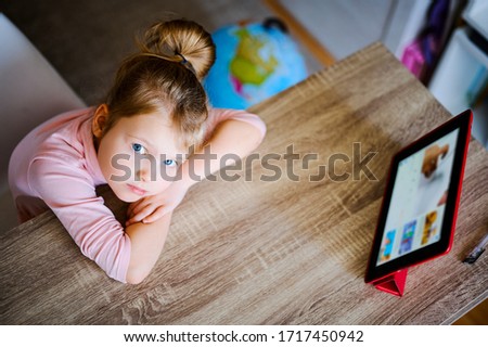 Young girl watches cartoons at home at a table on a digital tablet, a child plays with a device, a laptop computer