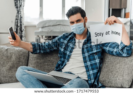 Man in medical mask holding smartphone, card with stay at home lettering and laptop on couch