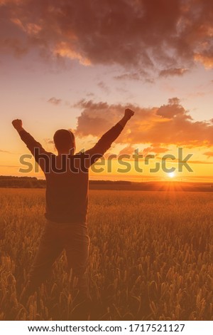 Silhouette of a man raise his hands up to sunset on the field with young rye or wheat in the summer with a cloudy sky background. Landscape.