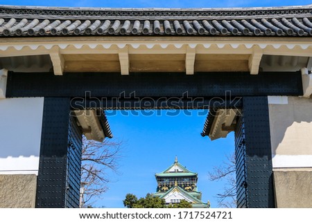 Japanese white grand imperial castle taken through a tradition door with blue sky background. Historical multiple stories white castle in a garden with big trees