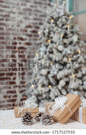 Square craft gifts boxes with snow-covered cones on table background of Christmas tree. Light brown cardboard boxes with white ribbons and bows against a brick wall and a snow-covered Christmas tree