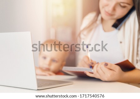 Young woman with baby speaks by phone and working with laptop on kitchen. Business Quarantine Concept.