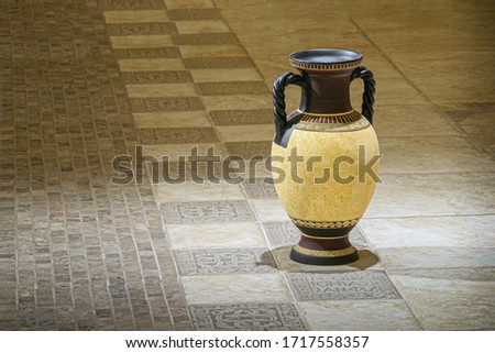 An elegant Greek amphora in beige is placed on colored terracotta in the room’s pasteurized beige tones. Amphora with two black handles with Greek ornament. The handles have a beautifully woven struct