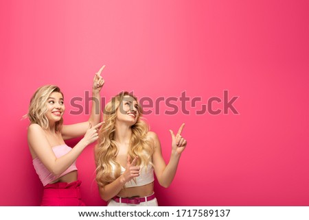Happy blonde girls pointing with fingers and looking up on pink background