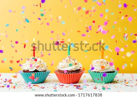 Three colorful  birthday cupcakes with candles and confetti on a yellow background