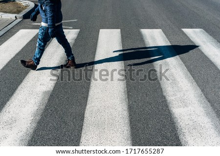 Present day man crosses the pedestrian crossing respecting the indicative traffic signs. The boy wears jeans, brown leather boots and a blue winter jacket to protect him from the cold