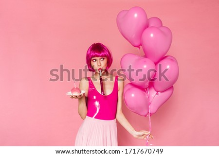 Image of amusing nice woman blowing in party horn while holding balloons and cake isolated over pink background