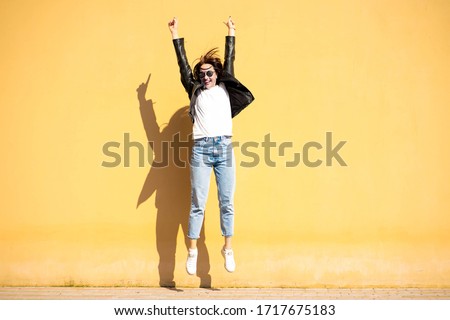 Brunette girl in a sunglasses and a casual clothing (white t shirt, blue jeans and a black jacket), is jumping and smiling in front of yellow wall. Picture with a colorful background and a copy space