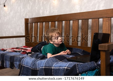 the child lies on a wooden sofa hiding in a blanket and looks at the laptop. rest at home.