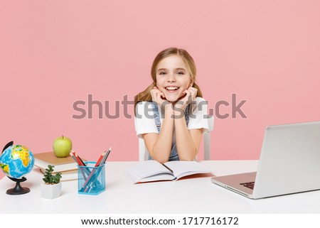 Smiling little kid schoolgirl 12-13 years old study at white desk with pc laptop isolated on pink background. School distance education at home during quarantine concept. Put hands prop up on chin