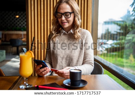 Elegant blonde woman sitting at cafe holding mobile phone and credit card signing up on website. Businesswoman paying with credit card while shopping online using mobile.