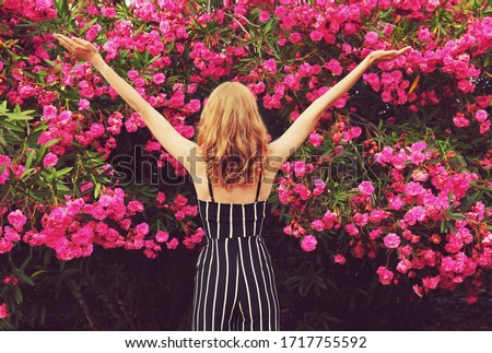 Happy young woman raises hands up over pink roses flowers background, view from the back