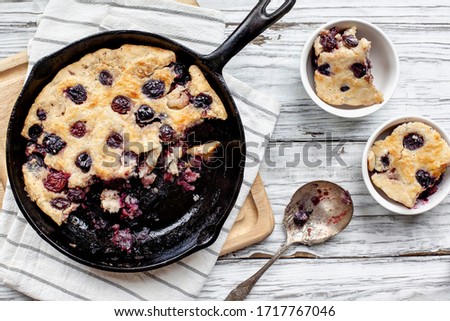 Fresh made homemade cherry cobbler baked in a cast iron pan with antique spoon and two servings over a rustic white wood table. Image shot from top view.