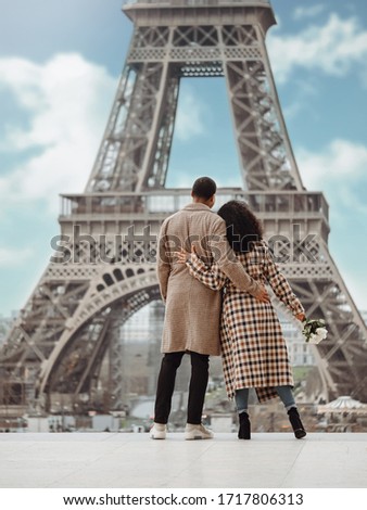 A young black couple, dressed in a plaid coat, stands in an embrace against the backdrop of the Eiffel Tower in Paris. Royalty-Free Stock Photo #1717806313
