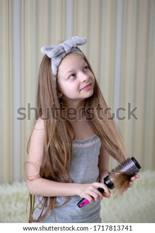 A girl with long blond hair sits and looks up thoughtfully, holding a hairbrash in her hands