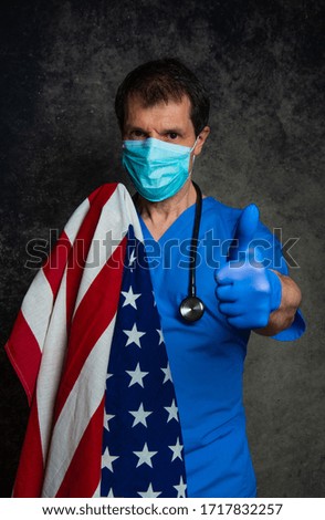 Doctor doing a thumb up hand sign, in blue hospital scrubs with face mask and stethoscope, with the American flag around his shoulders, against a dark studio background.