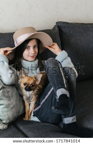 Charming girl of European race 8 years old. She wears a beige hat and smiles while sitting on the sofa with a chihuahua dog.