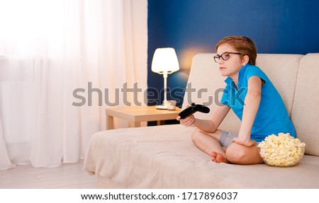 Blonde 9-year-old boy in a blue T-shirt and big glasses sitting on the couch and playing at home with a gamepad. Blue background and space for text