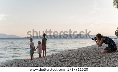 Young mother taking photos of her three kids, two boys and a girl, as they stand on the beach in the evening holding hands.