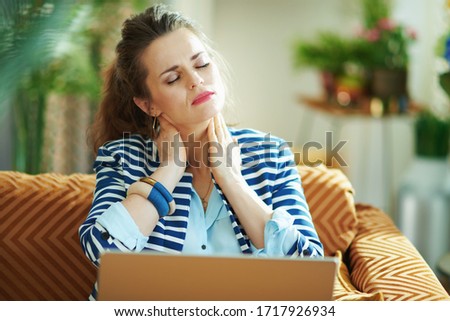 stressed stylish woman in blue blouse and striped jacket having neck pain sitting on couch with laptop at modern home in sunny day.