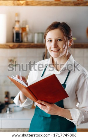 Portrait of young woman standing in the kitchen with recipe book in her hands. She looking into the camera and smile. Cooking at home concept, lifestyle. Vertical picture