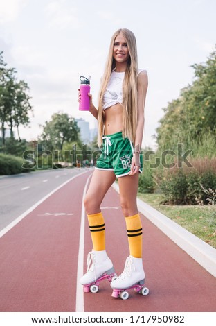 Very Beautiful young woman with long hair in vintage roller skates, green shorts and a white top, posing with a bottle of water. Summer evening on the street of a park.