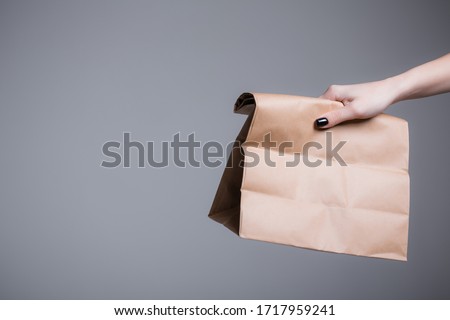 A female hand holds out or picks up a paper bag. The concept of food delivery and other things during quarantine. Gray background Royalty-Free Stock Photo #1717959241